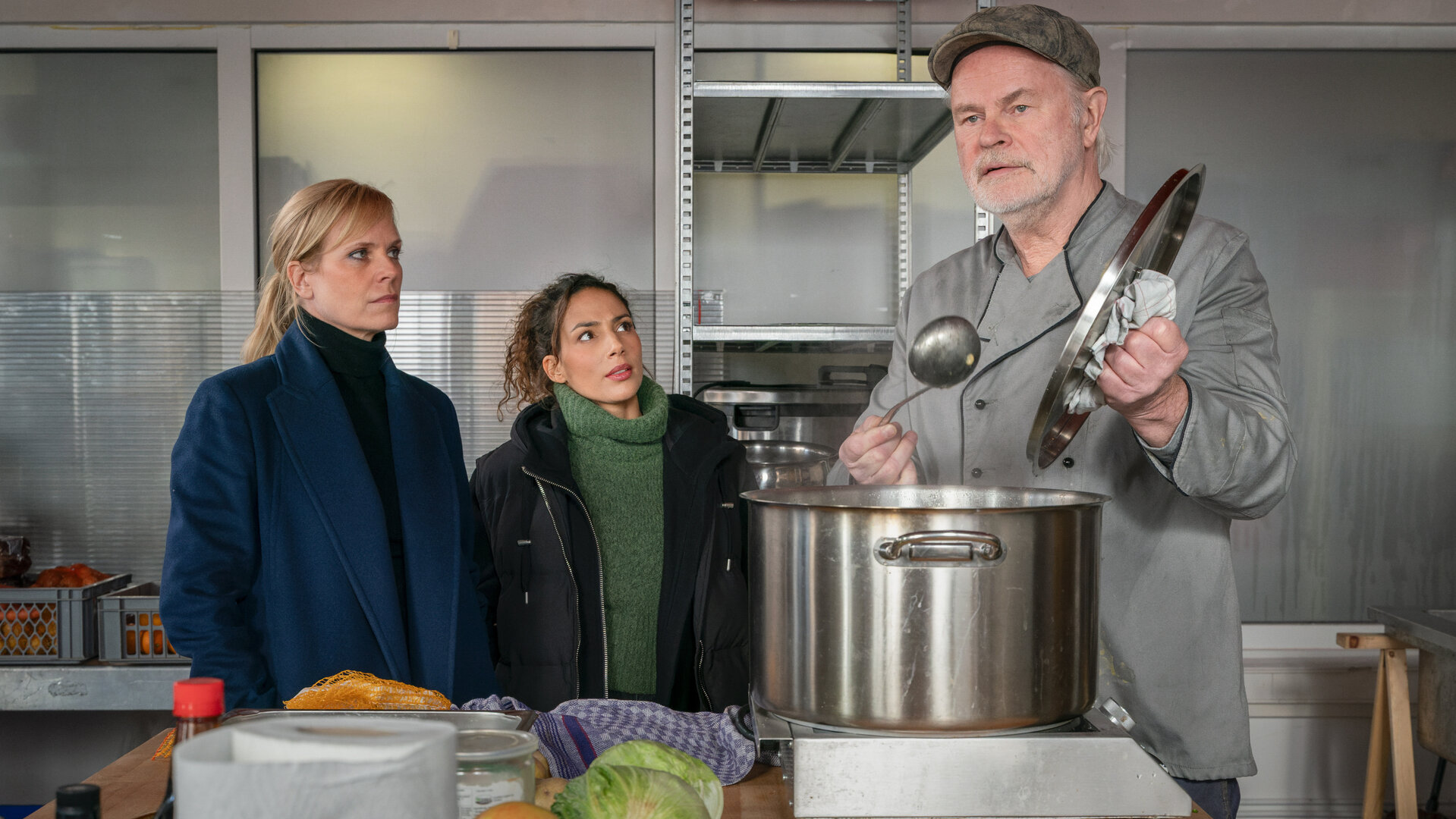 "SOKO Leipzig - Widerstand statt Ruhestand": Ina (Melanie Marschke) und Kim (Amy Mußul) befragen Klemenz Hill (Sebastian Kowski) in der Suppenküche. Klemenz steht vor einem großen Topf und hält in der einen Hand eine Suppenkelle und in der anderen den Topfdeckel.