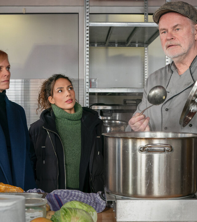 "SOKO Leipzig - Widerstand statt Ruhestand": Ina (Melanie Marschke) und Kim (Amy Mußul) befragen Klemenz Hill (Sebastian Kowski) in der Suppenküche. Klemenz steht vor einem großen Topf und hält in der einen Hand eine Suppenkelle und in der anderen den Topfdeckel.