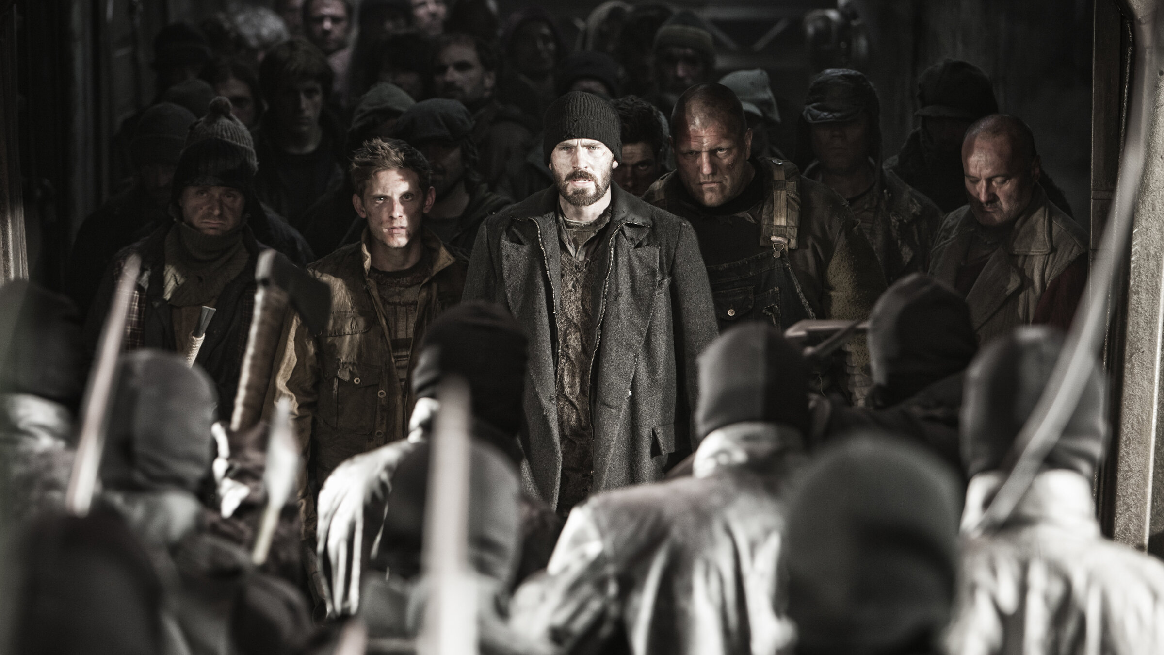 "Snowpiercer": Curtis (Chris Evans) und Edgar (Jamie Bell) stehen mit entschlossenem Blick in abgerissener Kleidung in einem Raum Gegnern gegenüber, die Sturmhauben tragen und mit Beilen bewaffnet sind. Diese Gegner sind unscharf im Vordergrund von hinten zu sehen. Neben und hinter Curtis und Edgar wiederum haben sich Männern aufgereiht, die wie die beiden einen verwahrlosten Eindruck machen. Sie sind unscharf im Halbdunkel zu erkennen.