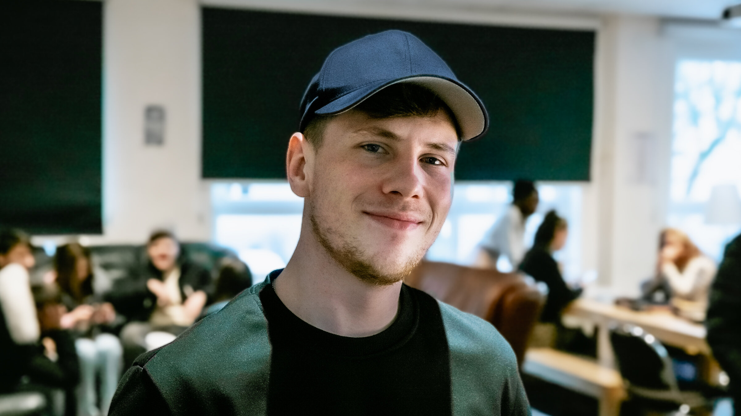 "37°: Vom Straßenjungen zum Streetworker": Pascal (24) steht im Aufenthaltsraum der Arche Hellersdorf, trägt eine blaue Baseballkappe, lächelt und blickt in die Kamera. Hinter ihm, unscharf zu erkennen, sitzen Jugendliche.