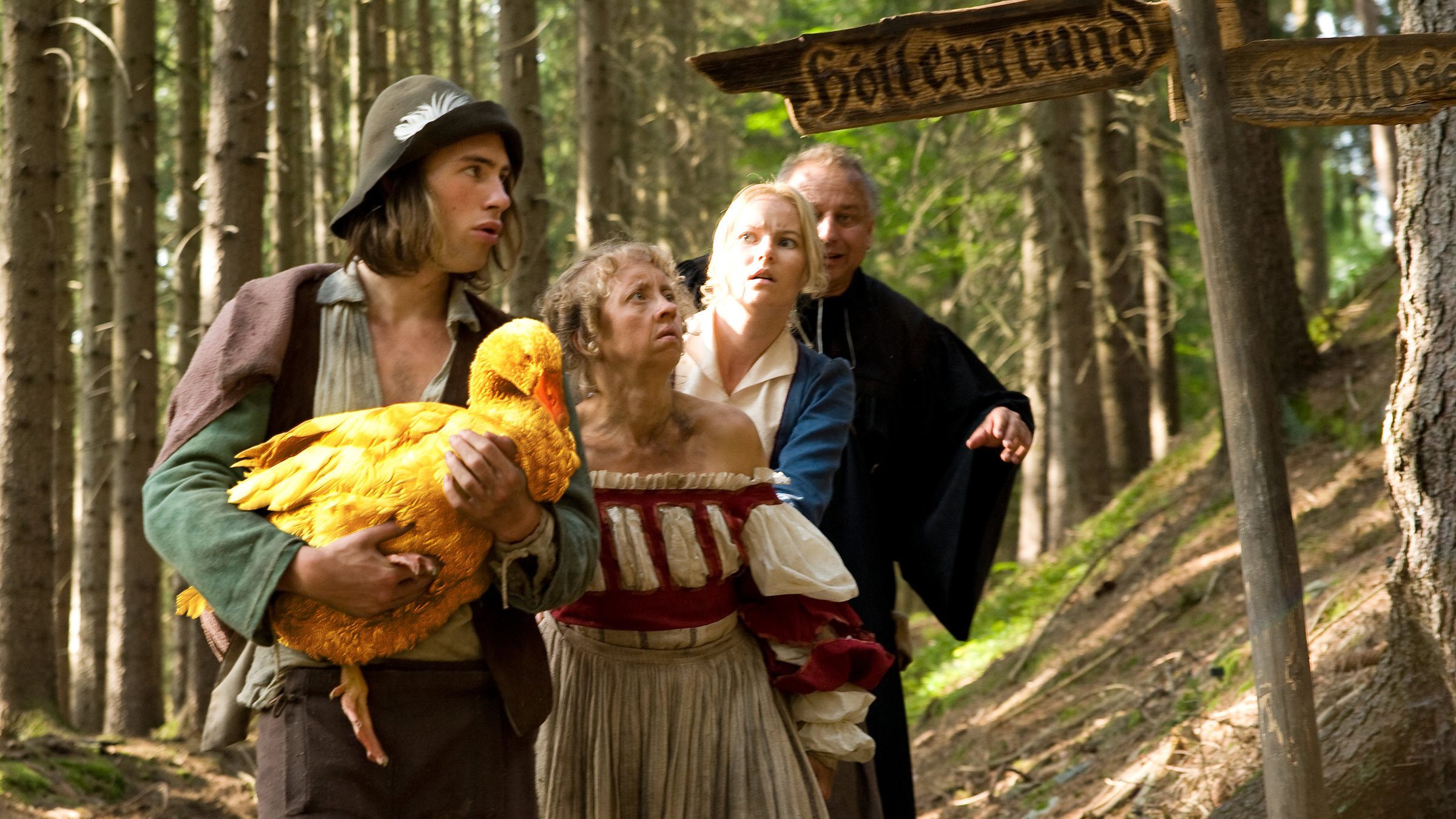 "Die Goldene Gans": Es ist Tag. Mitten im Wald steht der Dummling (Jeremy Mockridge) mit der Goldenen Gans in den Händen an einem Wegkreuz. Hinter ihm stehen die Wirtstöchter Liesel (Anna Böttcher) und Lotte (Teresa Weißbach) sowie der Pfarrer (Hilmar Eichhorn) in einer Reihe.