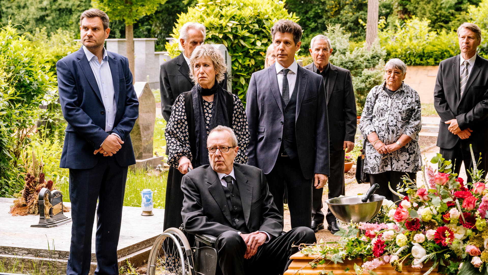 "SOKO Köln - Ruhe sanft": Dr. Philip Kraft (Thomas Clemens) steht mit seiner Familie auf dem Friedhof. Links im Bild steht sein Kollege Jonas Fischer (Lukas Piloty), der die Hände vor dem Körper verschränkt hat und ernst nach links blickt. Marianne Kraft (Beatrice Richter), die Mutter von Philip, steht neben ihrem Sohn und hält den Rollstuhl seines Onkels Holger (Dominik Bender) fest, der vor ihr sitzt. Die beiden blicken ebenfalls mit bedrückten Gesichtern nach links. Die weiteren Gäste der Beerdigung (Komparsen) und Philip sehen in die gleiche Richtung. Rechts vorne im Bild sieht man weiße, rote und rosa Blumen auf einem hölzernen Sarg liegen.
