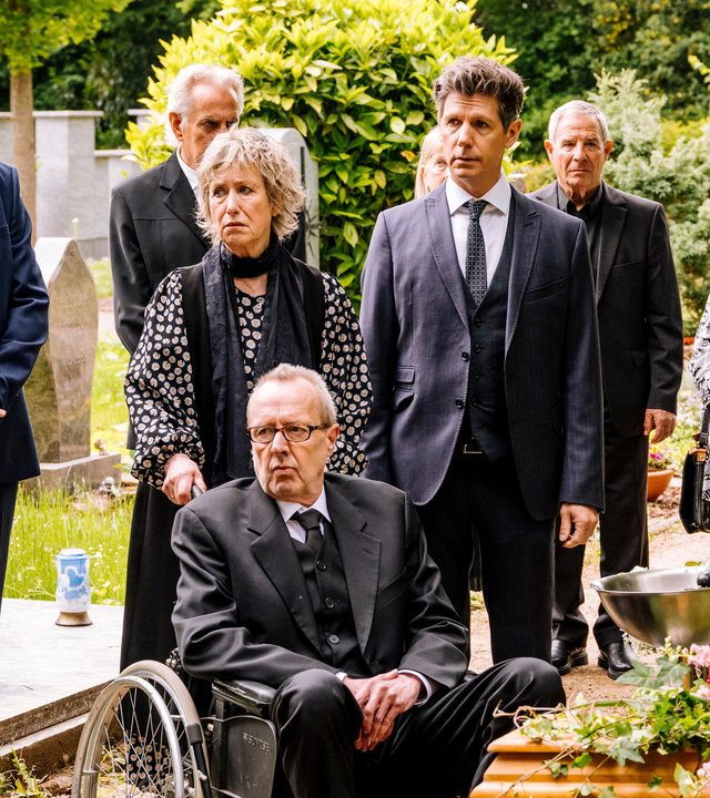 "SOKO Köln - Ruhe sanft": Dr. Philip Kraft (Thomas Clemens) steht mit seiner Familie auf dem Friedhof. Links im Bild steht sein Kollege Jonas Fischer (Lukas Piloty), der die Hände vor dem Körper verschränkt hat und ernst nach links blickt. Marianne Kraft (Beatrice Richter), die Mutter von Philip, steht neben ihrem Sohn und hält den Rollstuhl seines Onkels Holger (Dominik Bender) fest, der vor ihr sitzt. Die beiden blicken ebenfalls mit bedrückten Gesichtern nach links. Die weiteren Gäste der Beerdigung (Komparsen) und Philip sehen in die gleiche Richtung. Rechts vorne im Bild sieht man weiße, rote und rosa Blumen auf einem hölzernen Sarg liegen.