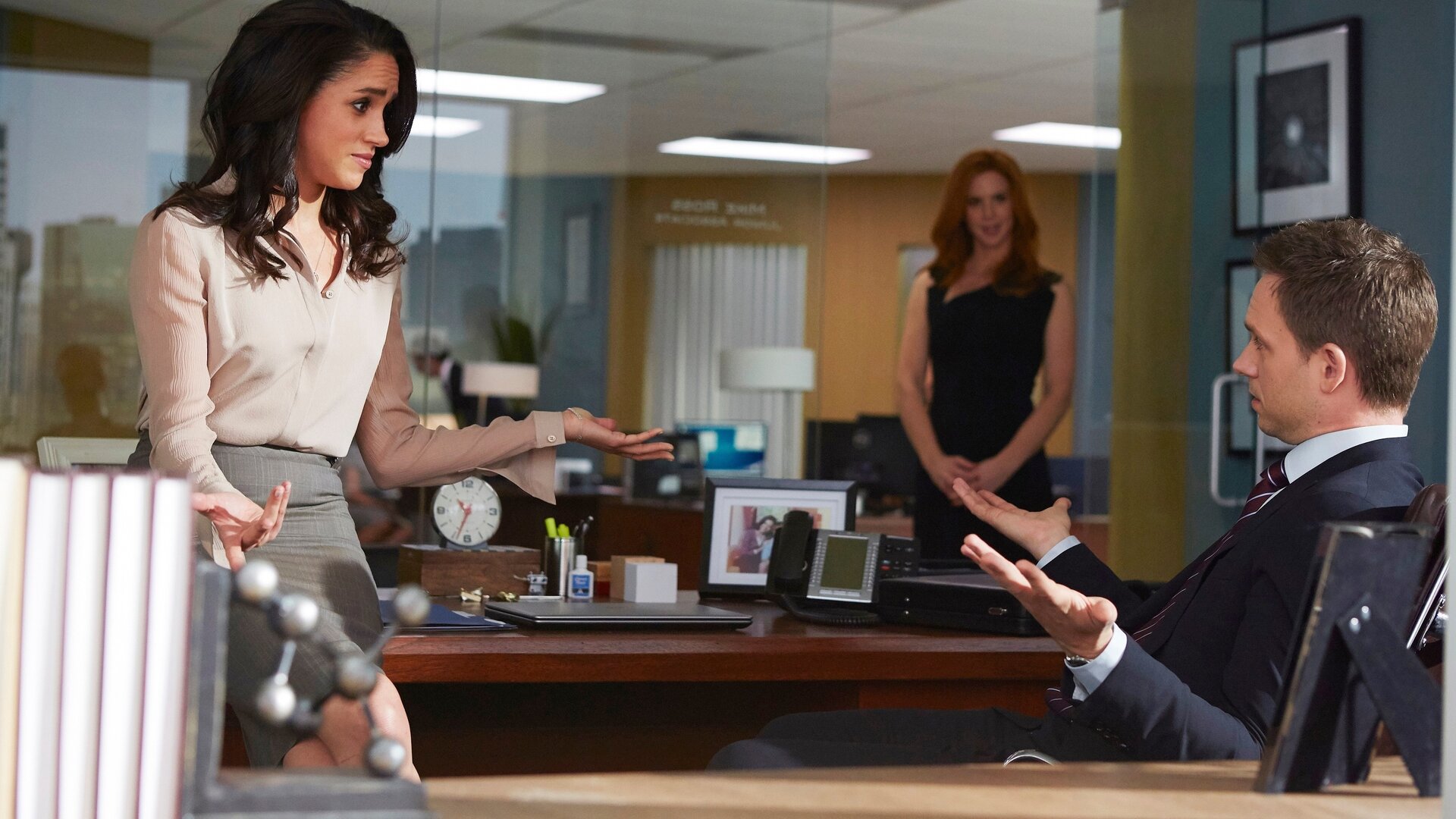 "Suits - Kämpfen, nicht kuscheln": Mike (Patrick J. Adams) sitzt auf einem Schreibtischstuhl, Rachel sitzt ihm zugewandt auf der Kante der Schreibtischplatte. Die beiden führen ein angeregtes Gespräch und gestikulieren mit ihren Händen, während Donna im Hintergrund steht und die Diskussion verfolgt.