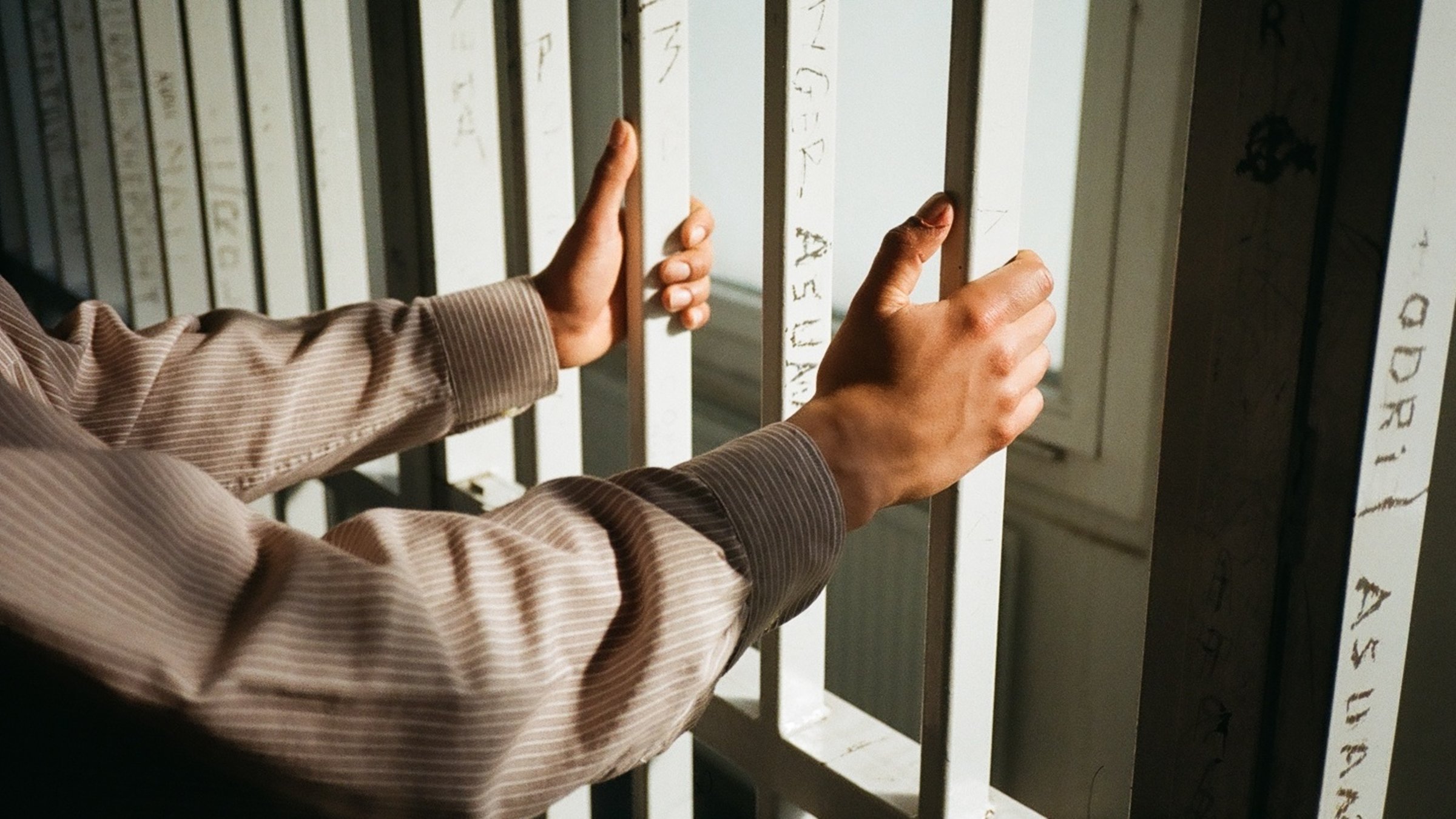 "KlassiXS: Macht und Hierarchien - Ein Mann steht hinter Gittern und hält zwei Gitterstäbe in der Hand. Er trägt ein grau-weiß-gestreiftes langärmliges Hemd, es sind nur seine Arme und Hände zu sehen. Auf die Gitterstäbe, die von dem Mann umfasst werden, sind Wörter und Namen eingeritzt.