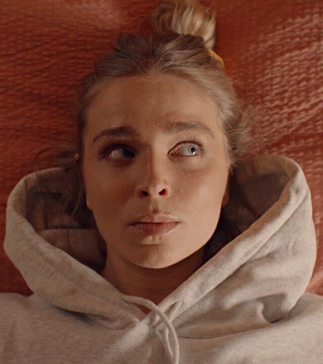 "Single Bells - 12 Dates till Christmas: Jens II": Blick von oben: Mina (Julie Van den Steen) liegt mit einem hellgrauen Hoodie bekleidet auf einem Kissen mit orangefarbenem Bezug.