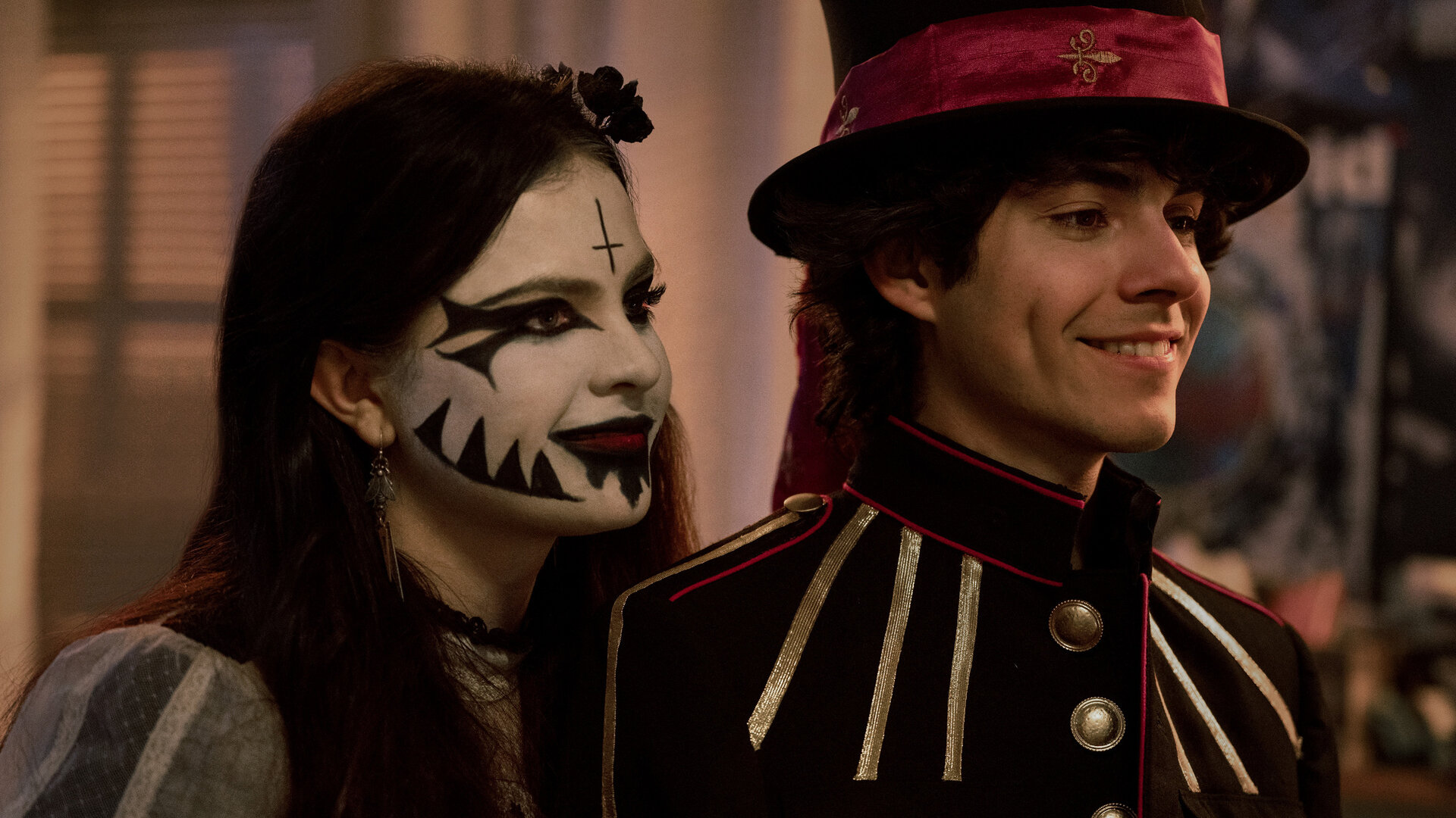 "Hysteria! - Die Ankunft des Bösen": Jordy (Chiara Aurelia) und Dylan (Emjay Anthony) stehen nebeneinander und blicken lächelnd in dieselbe Richtung. Jordy trägt ein auffälliges Make-up mit schwarzem Muster und ein dunkles Outfit, während Dylan eine Uniformjacke mit Zierknöpfen und einen Zylinder mit rotem Band trägt.