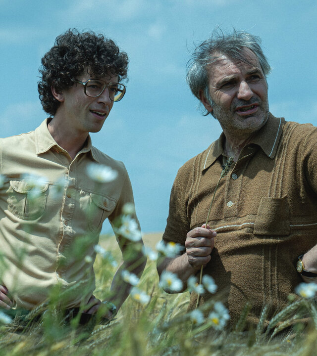 "We're on it, Comrades! - Lada und der Fluch der Hussiten": Vojta (Jan Cina) und David (Jiří Macháček) stehen zwischen Blumen und Weizenähren auf einem Feld.