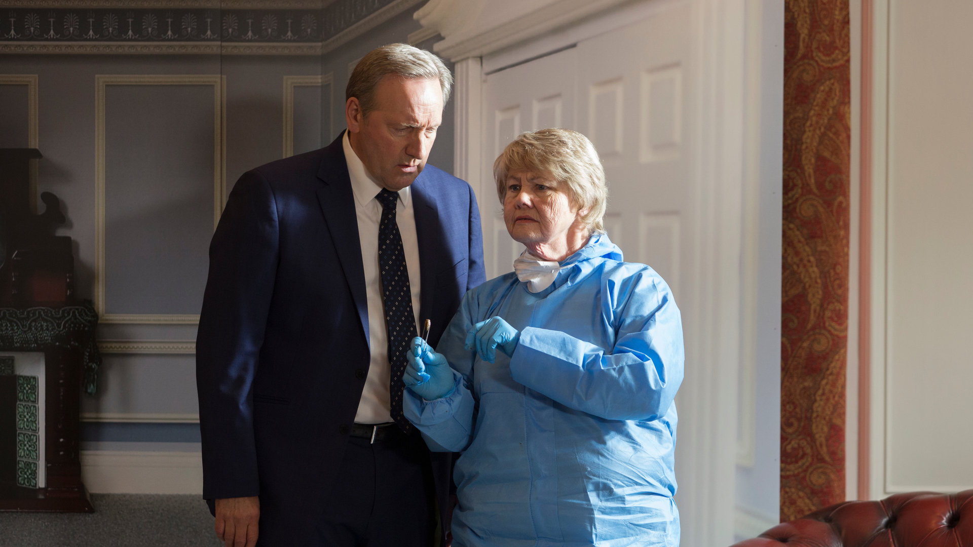 "Inspector Barnaby - Puppenmorde": Der mit einem dunklen Anzug bekleidete DCI John Barnaby (Neil Dudgeon) steht in einem Raum neben der einen hellblauen Schutzanzug und Latexhandschuhe tragenden Fleur Perkins (Annette Badland) in einem elegant vertäfelten Raum. Barnaby blickt Fleur konzentriert an, während sie nachdenklich in den Raum schaut.