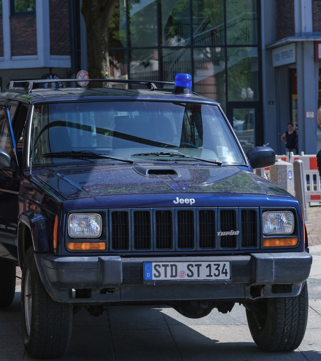 "SOKO Hamburg - Die erste Geige": Oskar Schütz (Marek Erhardt) rennt um einen schwarzen Jeep herum, sein Körper ist dabei nach links geneigt. Auf der anderen Seite des Autos schließt Franzi Berger (Paula Schramm) gerade die Beifahrertür. Auf dem Dach des Wagens befindet sich ein Blaulicht. Die Blicke der beiden Kommissare sind ernst. Das Auto steht auf einem gepflasterten Weg in der Stadt. Vor den Gebäuden im Hintergrund befinden sich rot-weiße Absperrungen.