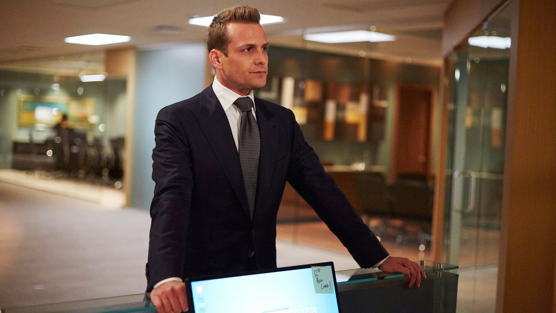 "Suits - Trennungsschmerzen": Harvey (Gabriel Macht) trägt einen dunklen Anzug mit Krawatte und steht im Flur der Anwaltskanzlei. Er hat seine Arme auf dem Tresen eines Arbeitsplatzes gestützt und schaut dabei nachdenklich.