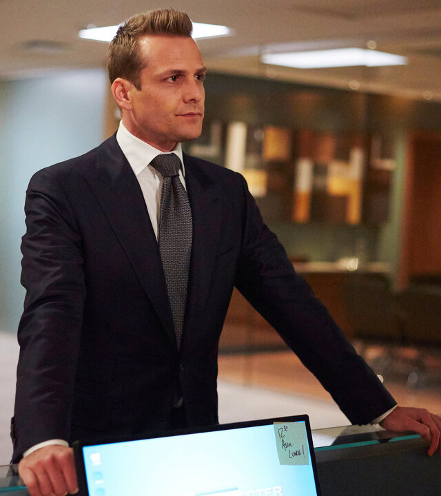 "Suits - Trennungsschmerzen": Harvey (Gabriel Macht) trägt einen dunklen Anzug mit Krawatte und steht im Flur der Anwaltskanzlei. Er hat seine Arme auf dem Tresen eines Arbeitsplatzes gestützt und schaut dabei nachdenklich.