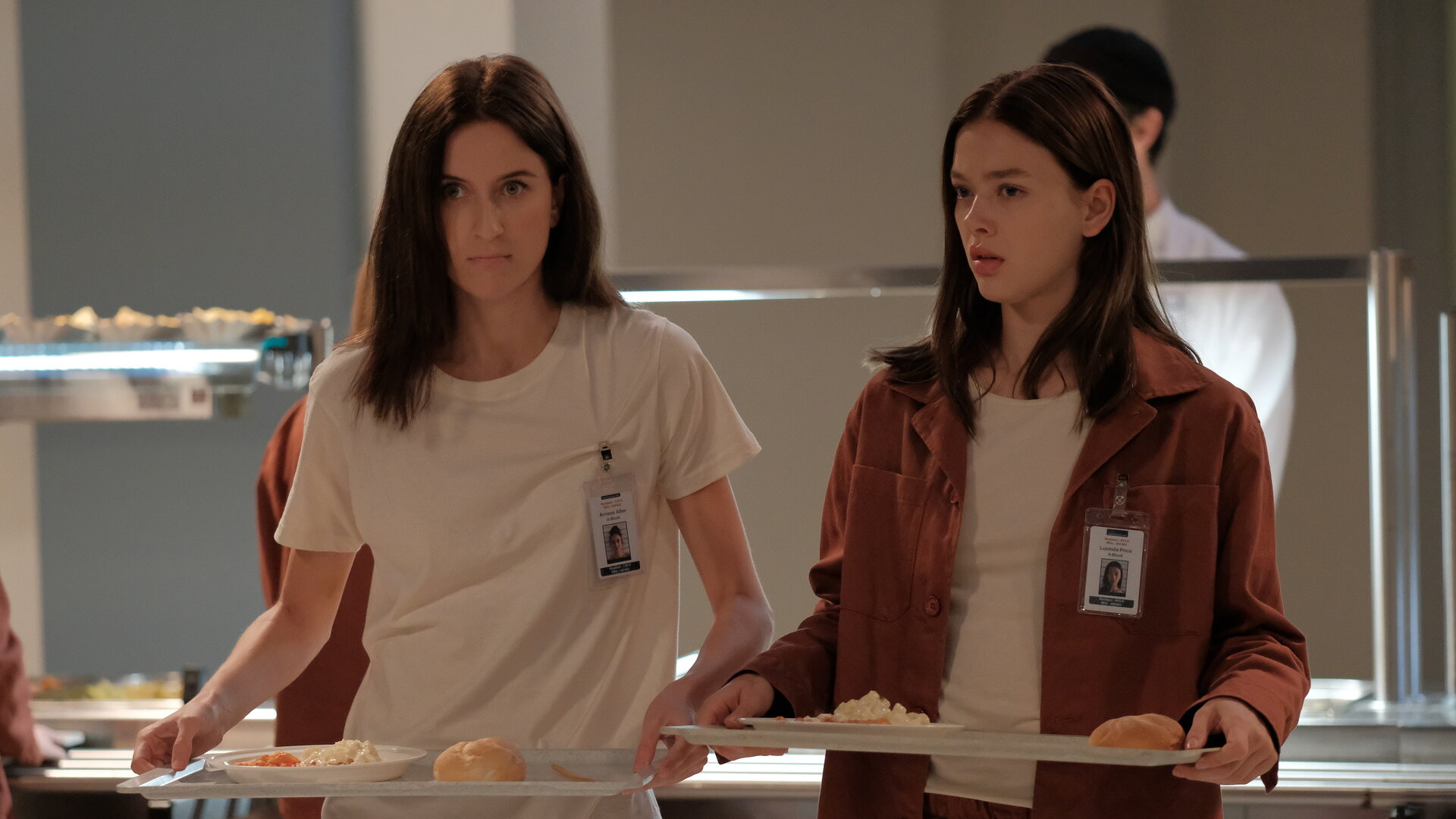 "Fallen - Eingewöhnung": Luce (Jessica Alexander) und Arriane (Josefine Koenig) stehen in der Kantine und tragen jeweils ein Tablett mit Essen. Beide blicken ernst, während hinter ihnen ein Mitarbeiter (Komparse) an der Essensausgabe arbeitet.