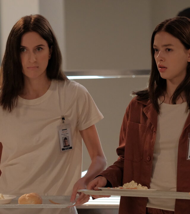 "Fallen - Eingewöhnung": Luce (Jessica Alexander) und Arriane (Josefine Koenig) stehen in der Kantine und tragen jeweils ein Tablett mit Essen. Beide blicken ernst, während hinter ihnen ein Mitarbeiter (Komparse) an der Essensausgabe arbeitet.