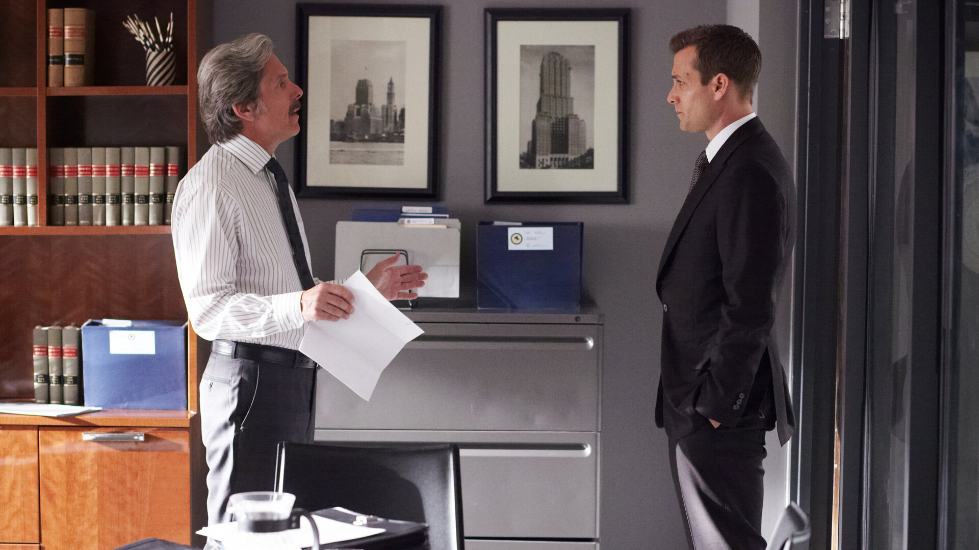 "Suits - Zweifelhafte Absichten": Harvey (Gabriel Macht) steht in einem hellen, modern eingerichteten Büro und blickt Cameron Dennis (Gary Cole) ernst an. Cameron hält ein Papier in der Hand und scheint im Gespräch mit Harvey zu argumentieren.