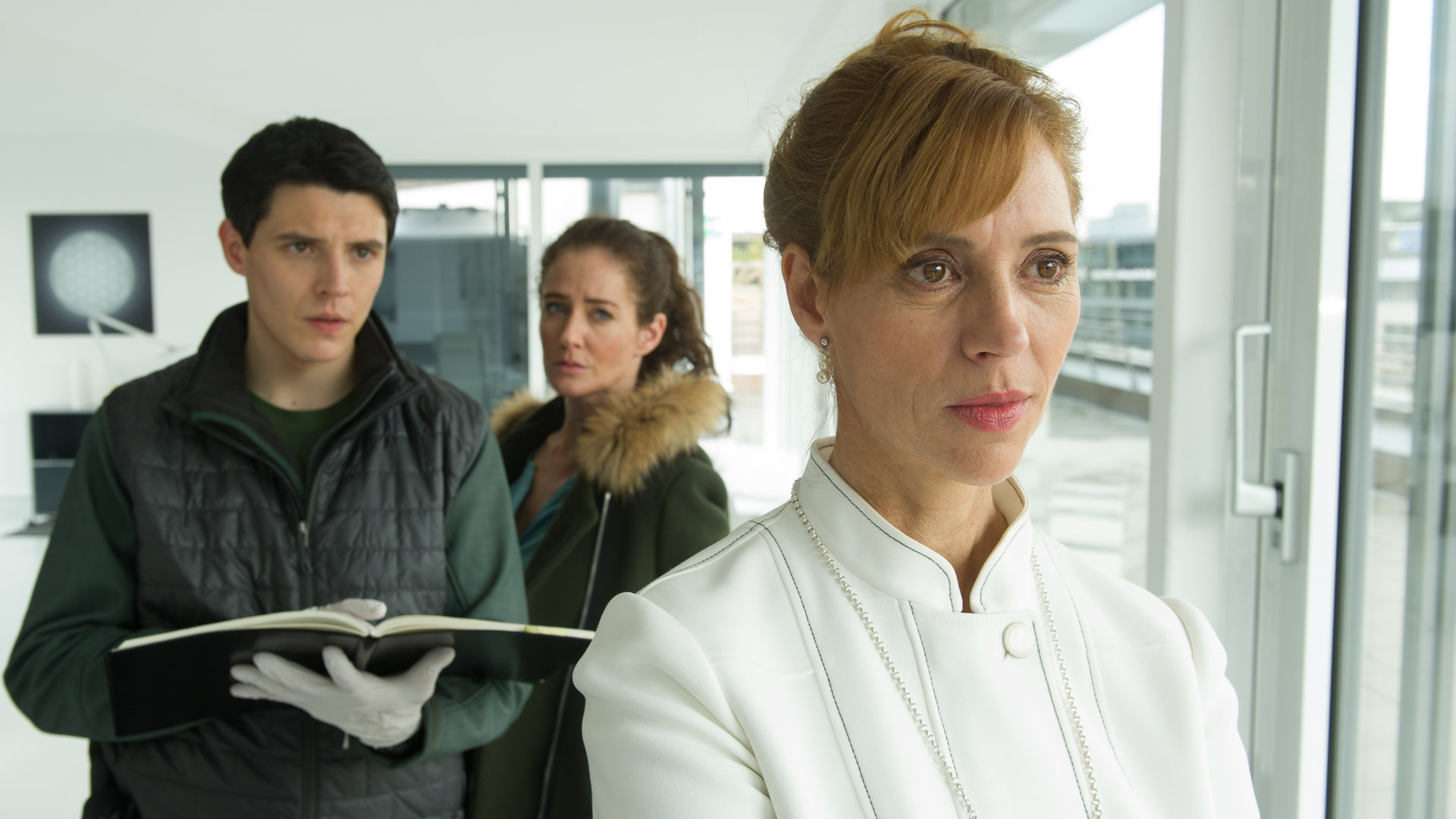 "SOKO Stuttgart - Wenn Sterne lügen": Selma Kirsch (Yve Burbach) und ihr Kollege Rico Sander (Benjamin Strecker) befragen die die Aura-Interpretin Chloe von Tretten (Carin C. Tietze) in deren modernen Praxisräumen über den Dächern von Stuttgart. Rico trägt Gummihandschuhe und hält ein aufgeschlagenes Terminbuch in den Händen.