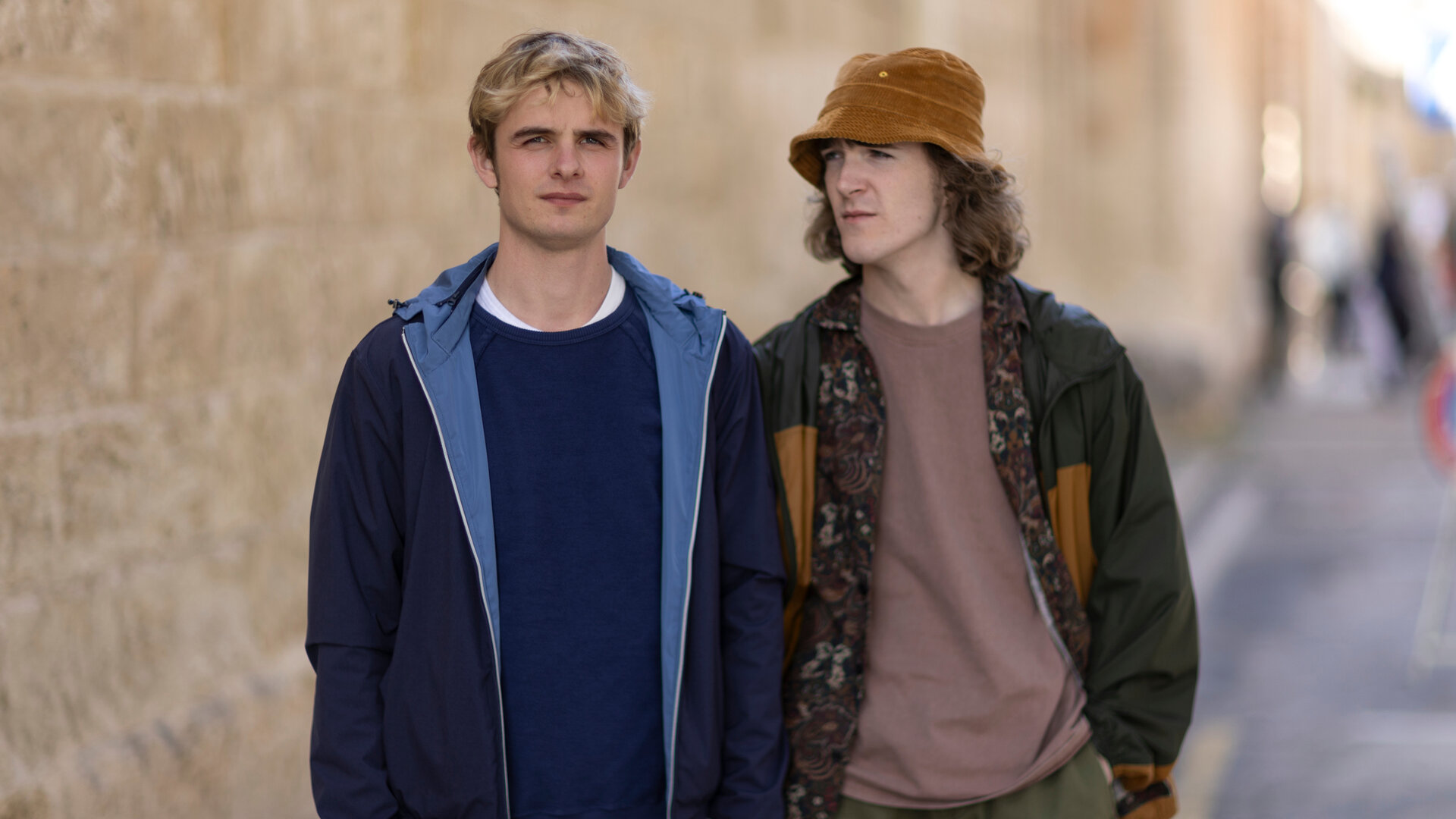 "Alex Rider - Labor": Alex (Otto Farrant) steht mit nachdenklichem Gesichtsausdruck vor einer sandfarbenen Steinmauer, während Tom (Brenock O'Connor) neben ihm steht und ihn von der Seite ansieht. Im Hintergrund sind unscharf Passanten zu erkennen.