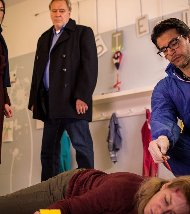 "SOKO München - Rabaukenhaus": Im Flur einer Kita liegt eine Frauenleiche auf dem Boden, Dr. Weissenböck (Florian Odendahl), sitz in der Hocke neben ihr und deutet mit einem Kugelschreiber auf ihren Hinterkopf. Katharina Hahn (Bianca Hein) und Arthur Bauer (Gerd Silberbauer) stehen daneben und schauen auf die Leiche.