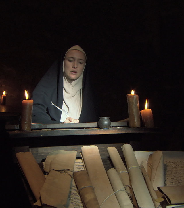 "Die glorreichen 10 - Die erfolgreichsten Mediziner der Geschichte": Darstellerin der Hildegard von Bingen sitzt in einem mit Kerzen beleuchteten Klosterraum an einem Pult und schreibt.