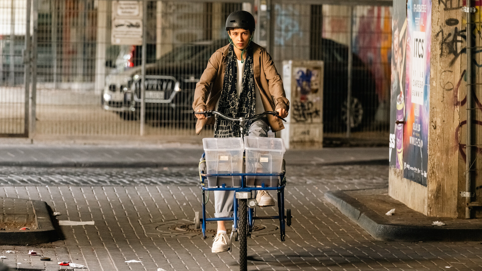 "SOKO Köln - Ming Pilz": Momo Malé (Shadi Eck) fährt auf einem Fahrrad durch eine verschmutze Unterführung. Vorne auf dem Fahrrad befinden sich zwei Plastikboxen mit braunem Inhalt. Er trägt einen Fahrradhelm und sieht ernst nach vorne.