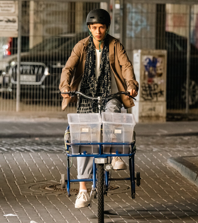 "SOKO Köln - Ming Pilz": Momo Malé (Shadi Eck) fährt auf einem Fahrrad durch eine verschmutze Unterführung. Vorne auf dem Fahrrad befinden sich zwei Plastikboxen mit braunem Inhalt. Er trägt einen Fahrradhelm und sieht ernst nach vorne.