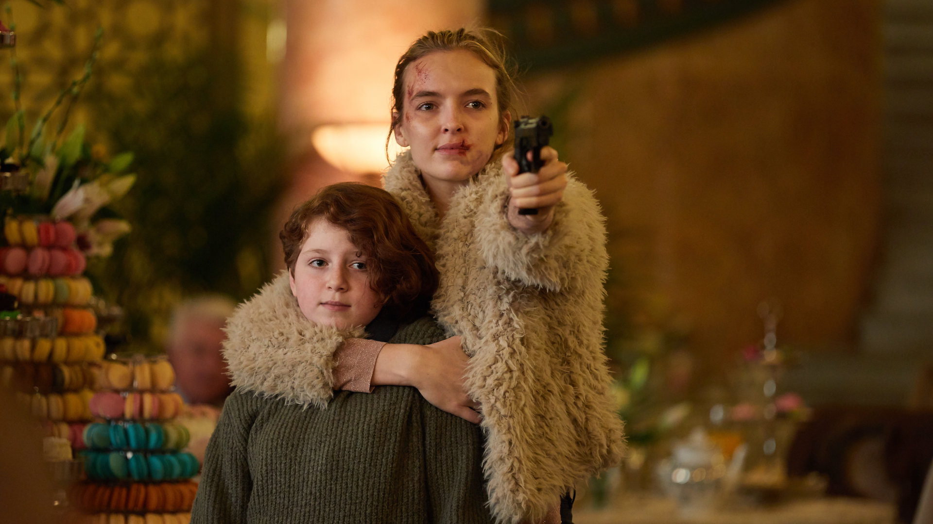 "Killing Eve - Gott, bin ich müde": Villanelle (Jodie Comer) hält Irina (Yuli Lagodinsky) in einem schicken Restaurant nah bei sich und zielt gleichzeitig mit einer Pistole nach vorne.