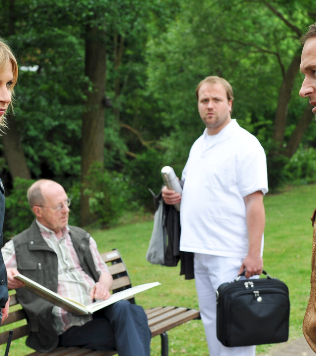 "Notruf Hafenkante - Kais Entscheidung": Melanie (Sanna Englund) und Kai (Markus Knüfken) stehen voreinander in einem Garten und schauen sich ratlos an. Im Hintergrund steht der Pfleger Matthias Rehmert (Moritz Sachs) vor einer Bank, auf der Seniorenheimbewohner Müller (Hans Peter Korff) sitzt und in einem Buch blättert.