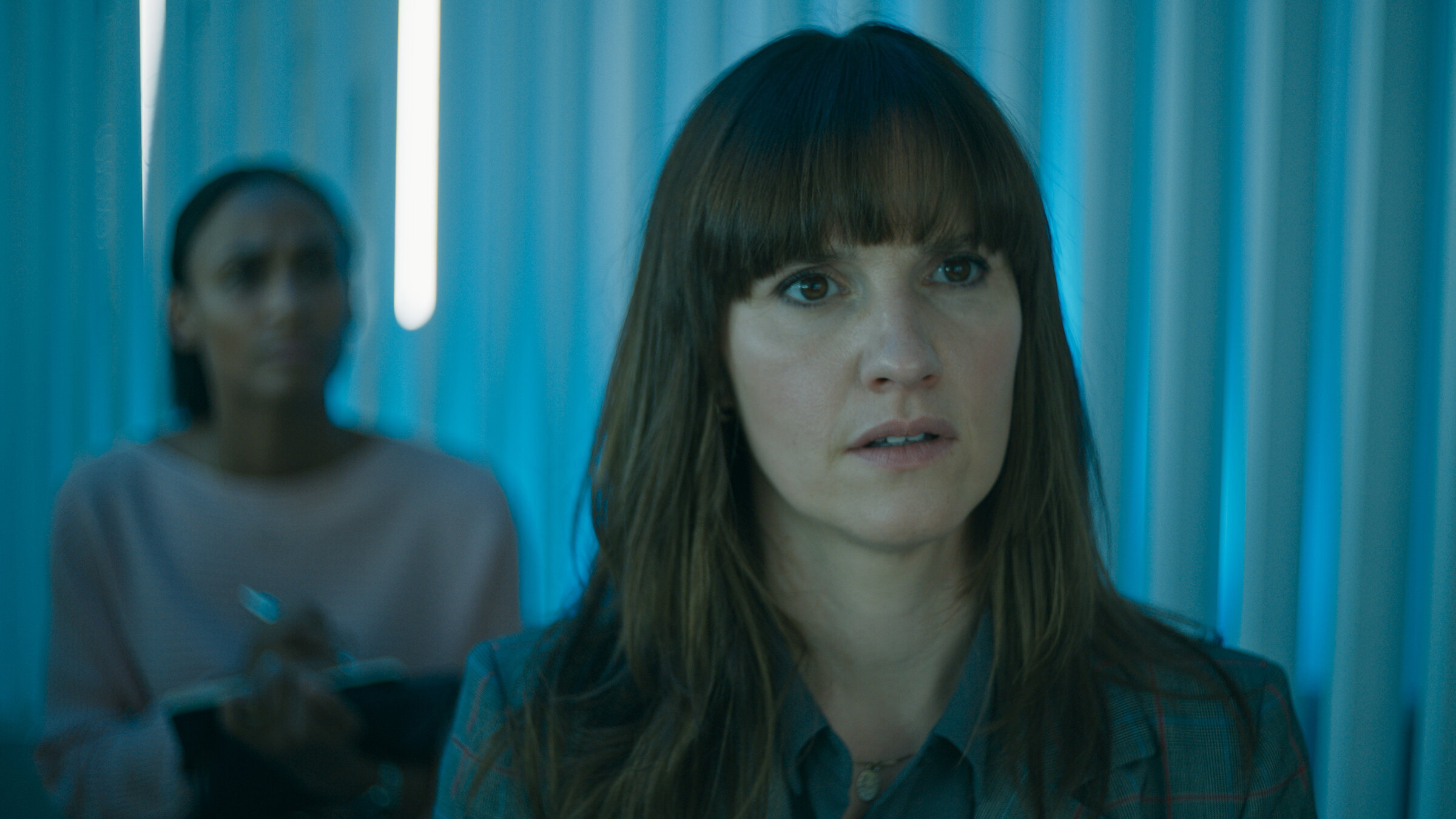 "Concordia - Tödliche Utopie (2) - Der Verdacht": Thea Ryan (Ruth Bradley) steht in einem Innenraum und schaut zur Seite. Hinter ihr steht, unscharf zu erkennen, Isabelle Larsson (Nanna Blondell).