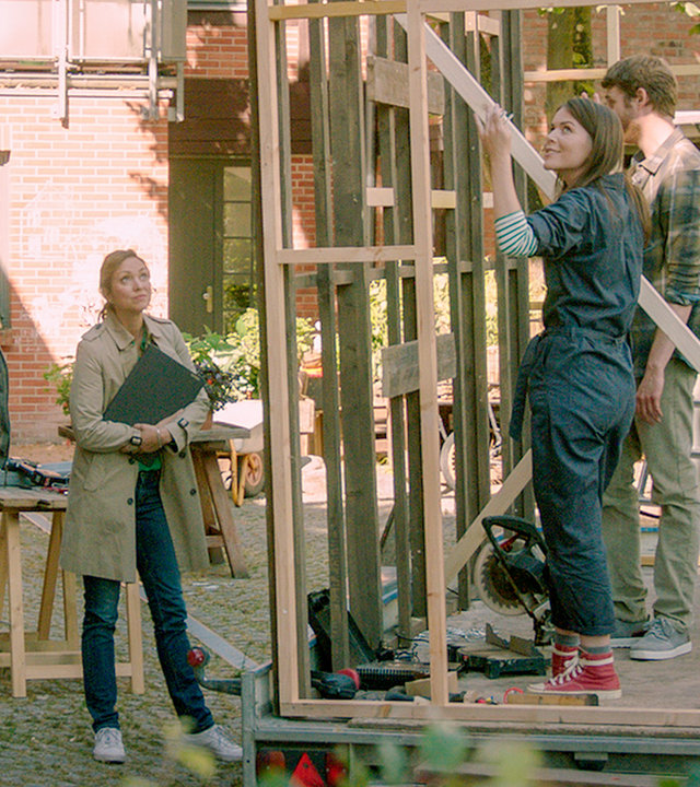 "SOKO Wismar – Ausgesorgt": Im Innenhof eines Mehrfamilienhauses steht ein sich im Bau befindliches "Tiny House". Nur die Form der Hütte ist bereits durch die Holzkonstruktion zu erkennen. In der Hütte stehen Alexandra Lämmle (Antonia Leichtle) und Florian Hein (Jonas Anders). Außerhalb und davor stehen Lars Pöhlmann (Dominic Boeer) und Karoline Joost (Nike Fuhrmann), die zu den beiden Personen in der Hütte blicken.