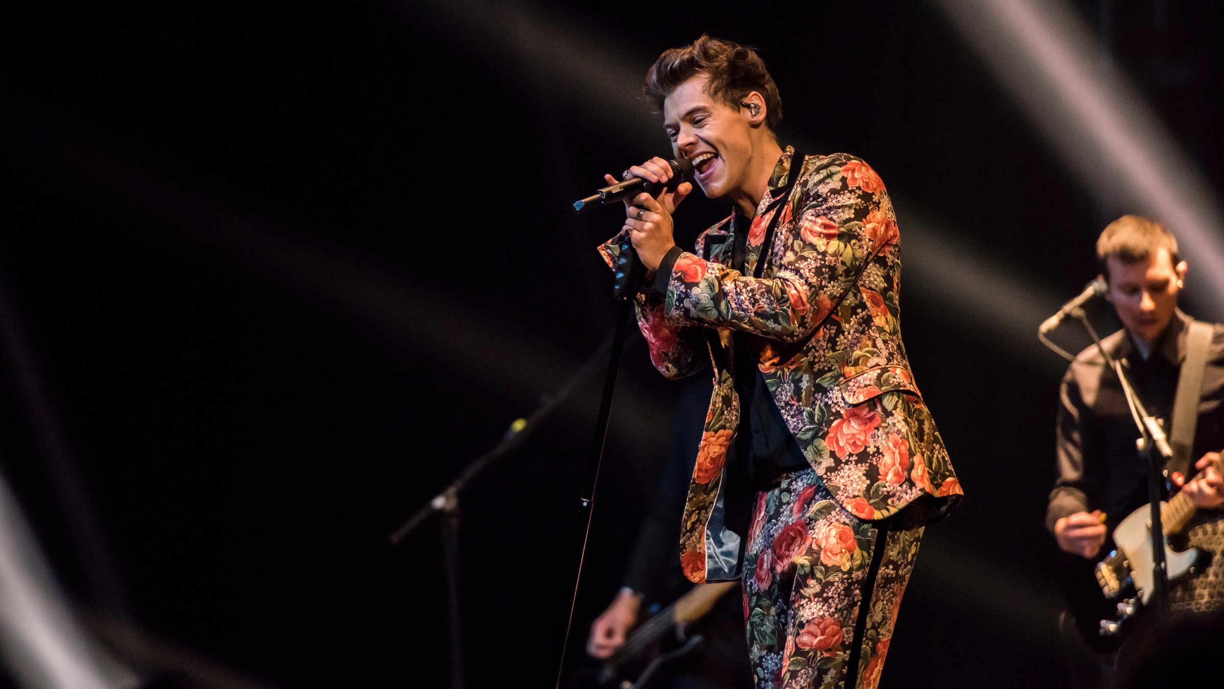 "Pop Around The Clock: Harry Styles: Live in Manchester": Ein junger Mann und einem bunten Anzug steht auf einer dunklen Bühne und singt in sein Mikrofon.