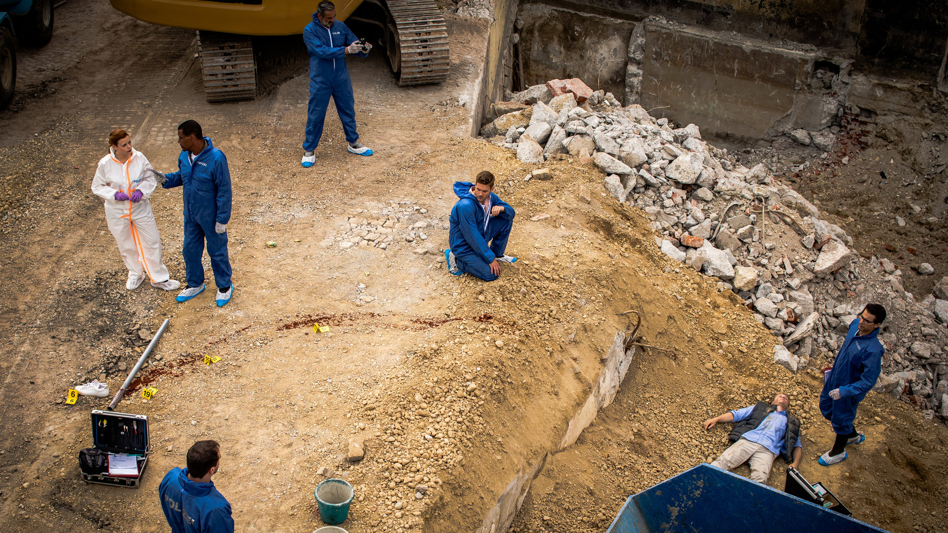 "SOKO München - Stille Liebe": Das Team der SOKO befindet sich am Tatort auf einer Baustelle. Dr. Weissenböck (Florian Odendahl) steht am Abhang der Baustelle neben der Leiche eines Mannes. Der Tote liegt auf dem Rücken vor einem blauen Container. Neben der Leiche steht ein Spurensicherungskoffer. Dominik Morgenstern (Joscha Kiefer) kniet in der Mitte des Platzes auf dem steinigen und sandigen Boden und blickt zu einem Kollegen, der neben einem weiteren Spurensicherungskoffer und einer blutigen Metallstange steht. Billie Curio (Sina Reiß) steht auf der linken Seite und unterhält sich mit Franz Ainfachnur (Christofer von Beau). Hinter ihnen steht ein weiterer Mitarbeiter und fotografiert den Tatort. Die Männer des Teams tragen blaue Schutzanzüge, sowie blau-weißen Schutzüberzüge für die Schuhe. Billie Curio trägt einen weißen Anzug, mit weißen Schutzüberzügen an den Füßen und lila Gummihandschuhen.