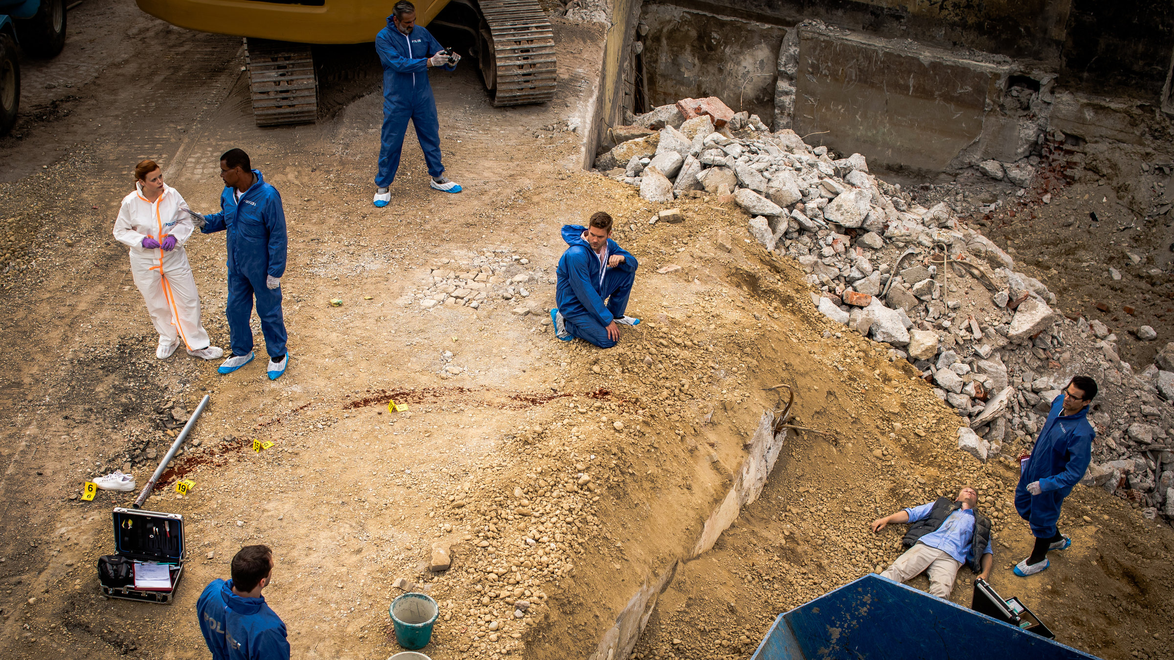 "SOKO München - Stille Liebe": Das Team der SOKO befindet sich am Tatort auf einer Baustelle. Dr. Weissenböck (Florian Odendahl) steht am Abhang der Baustelle neben der Leiche eines Mannes. Der Tote liegt auf dem Rücken vor einem blauen Container. Neben der Leiche steht ein Spurensicherungskoffer. Dominik Morgenstern (Joscha Kiefer) kniet in der Mitte des Platzes auf dem steinigen und sandigen Boden und blickt zu einem Kollegen, der neben einem weiteren Spurensicherungskoffer und einer blutigen Metallstange steht. Billie Curio (Sina Reiß) steht auf der linken Seite und unterhält sich mit Franz Ainfachnur (Christofer von Beau). Hinter ihnen steht ein weiterer Mitarbeiter und fotografiert den Tatort. Die Männer des Teams tragen blaue Schutzanzüge, sowie blau-weißen Schutzüberzüge für die Schuhe. Billie Curio trägt einen weißen Anzug, mit weißen Schutzüberzügen an den Füßen und lila Gummihandschuhen.