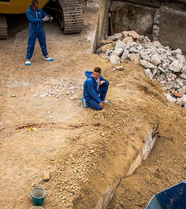 "SOKO München - Stille Liebe": Das Team der SOKO befindet sich am Tatort auf einer Baustelle. Dr. Weissenböck (Florian Odendahl) steht am Abhang der Baustelle neben der Leiche eines Mannes. Der Tote liegt auf dem Rücken vor einem blauen Container. Neben der Leiche steht ein Spurensicherungskoffer. Dominik Morgenstern (Joscha Kiefer) kniet in der Mitte des Platzes auf dem steinigen und sandigen Boden und blickt zu einem Kollegen, der neben einem weiteren Spurensicherungskoffer und einer blutigen Metallstange steht. Billie Curio (Sina Reiß) steht auf der linken Seite und unterhält sich mit Franz Ainfachnur (Christofer von Beau). Hinter ihnen steht ein weiterer Mitarbeiter und fotografiert den Tatort. Die Männer des Teams tragen blaue Schutzanzüge, sowie blau-weißen Schutzüberzüge für die Schuhe. Billie Curio trägt einen weißen Anzug, mit weißen Schutzüberzügen an den Füßen und lila Gummihandschuhen.