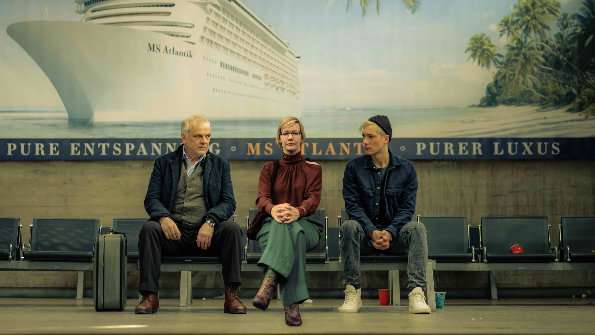 "Das schwarze Quadrat": Vincent Kowalski (Bernhard Schütz), Martha (Sandra Hüller) und Nils Forsberg (Jacob Matschenz) sitzen im Wartebereich eines Kreuzfahrtterminal nebeneinander. Hinter ihnen hängt ein großes Wandbild, das ein Kreuzfahrtschiff neben einem tropischen Sandstrand zeigt.
