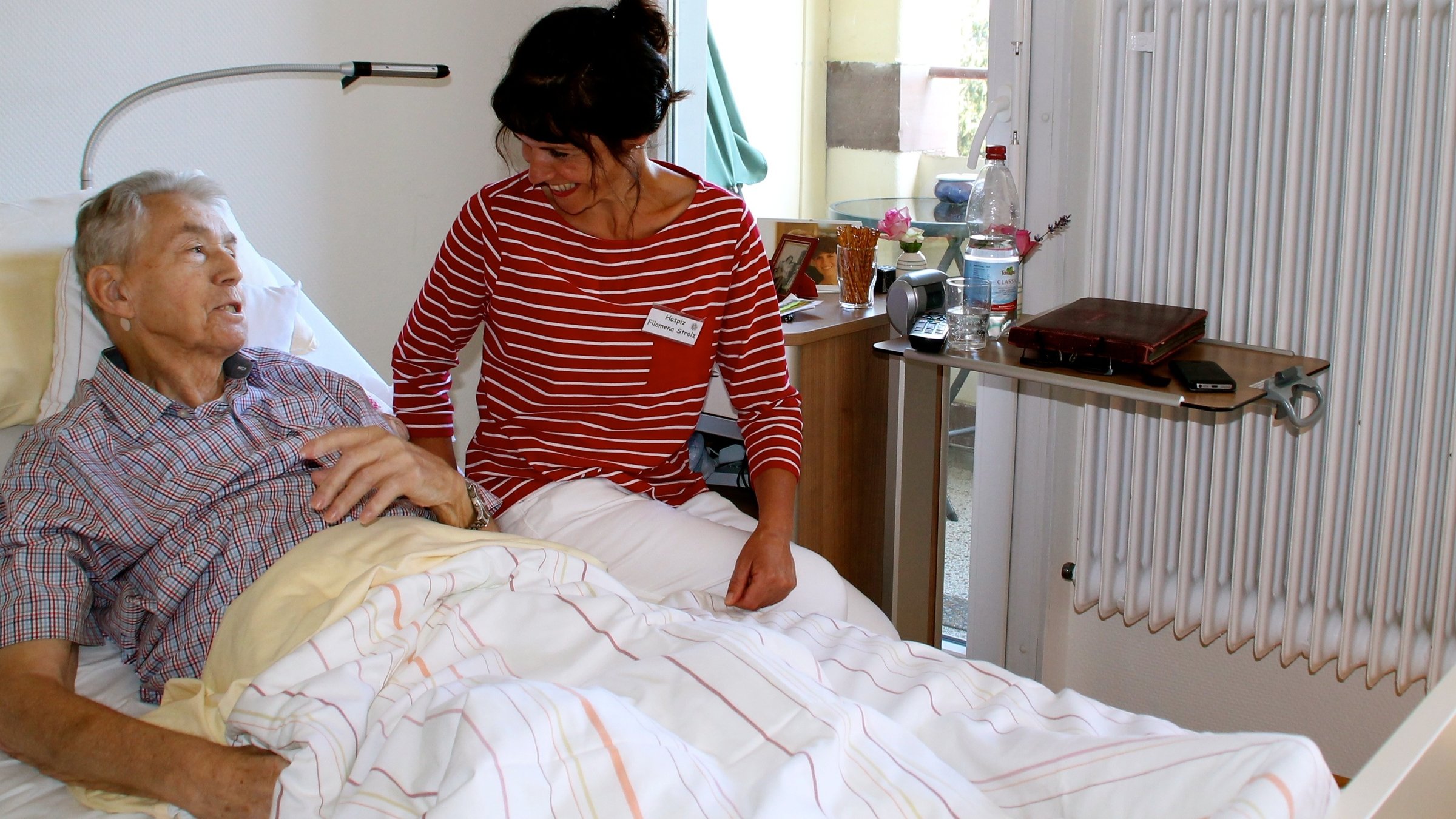 "37°: Der Geschmack von Leben - Die Köchin, das Hospiz und ein gutes Ende": Pflegerin Filomena steht neben Eduard Nöther am Krankenbett und lächelt ihm zu.