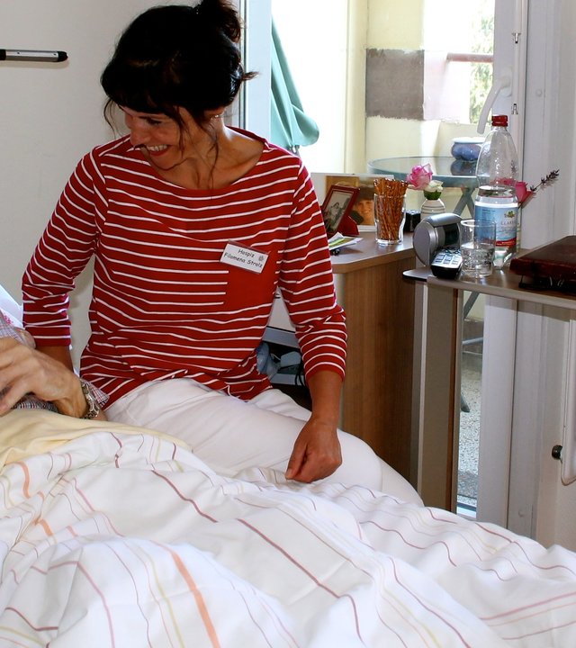 "37°: Der Geschmack von Leben - Die Köchin, das Hospiz und ein gutes Ende": Pflegerin Filomena steht neben Eduard Nöther am Krankenbett und lächelt ihm zu.