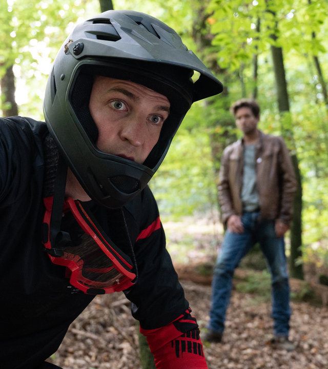 "SOKO Stuttgart - Trail des Todes": Jo Stoll (Peter Ketnath) und Philipp Bode (Jannik Mühlenweg) befinden sich im Wald. Bode, der im Vordergrund zu sehen ist, trägt Fahrradfunktionskleidung, einen Fahrradhelm mit Kinnschutz und eine Schutzbrille, die ihm um den Hals hängt. Es ist anzunehmen, dass er auf seinem Fahrrad sitzt. Sein Gesichtsausdruck wirkt geschockt, er hat die Augen weit aufgerissen. Jo ist leicht unscharf zu erkennen und steht mit etwas Abstand hinter ihm.