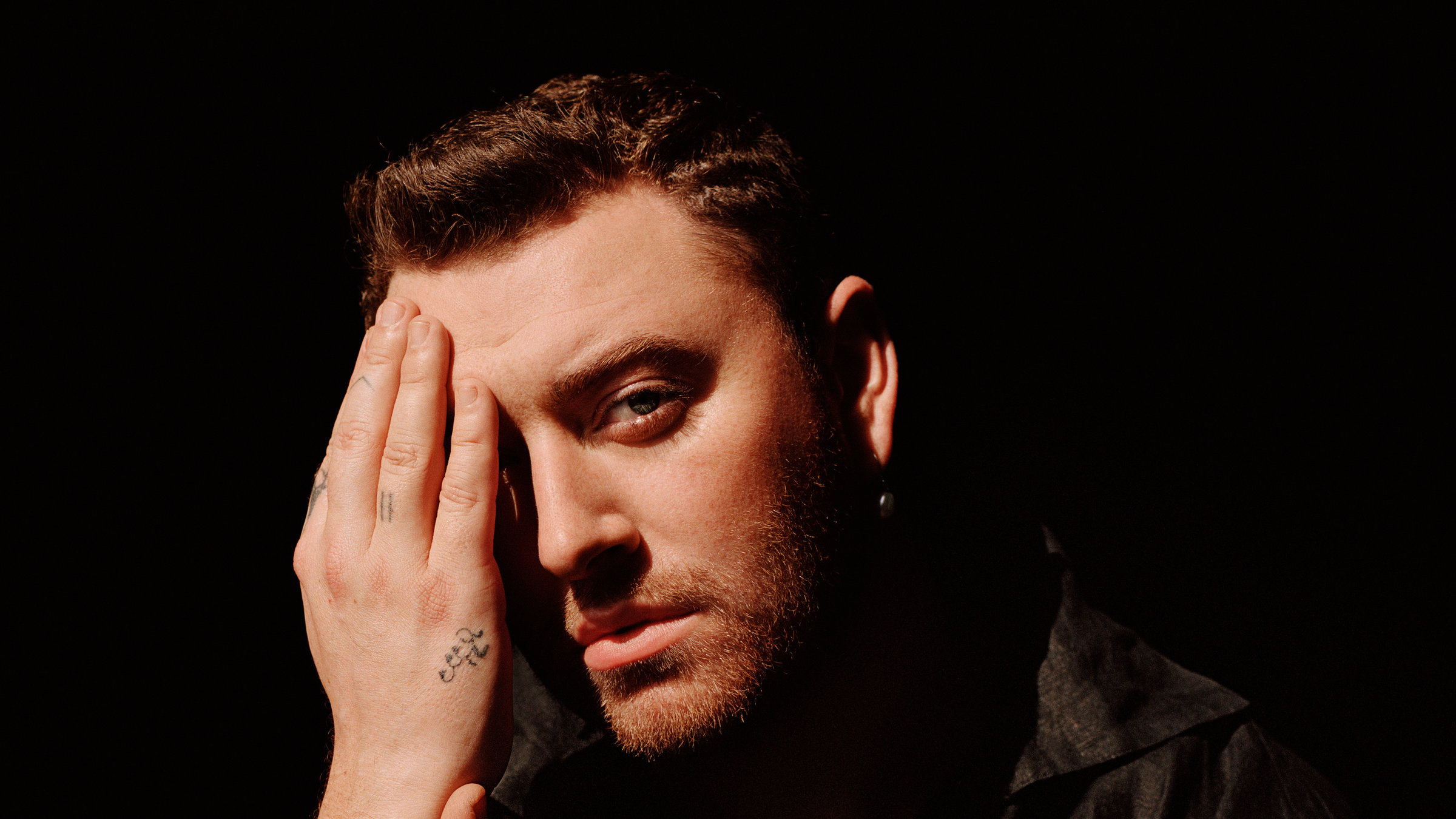 "Pop Around the Clock: Sam Smith: Live at the Royal Albert Hall": Ein Mann mit kurzen dunklen Haaren sitzt vor einem schwarzen Hintergrund mit einem schwarzen Hemd. Einen Arm hat er aufgestützt, mit der anderen Hand bedeckt er die Hälfte seines Gesichts.