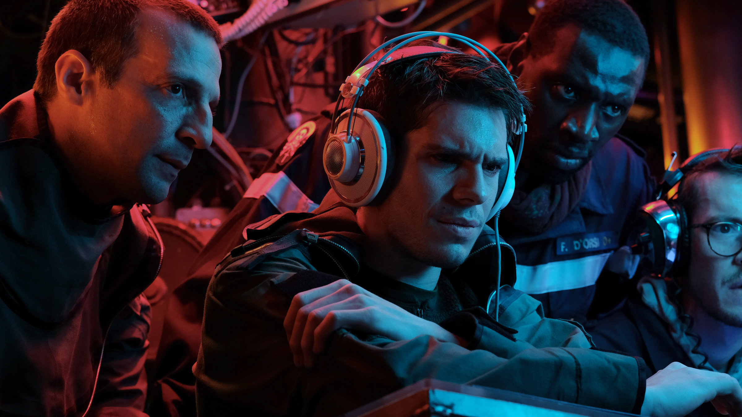 "Entscheidung in der Tiefe": Admiral FOST (Mathieu Kassovitz) und D’Orsi (Omar Sy) blicken auf einen nicht sichtbaren Bildschirm. Vor ihnen sitzt der Chanteraide (François Civil) mit konzentrierter Miene und Kopfhörern.