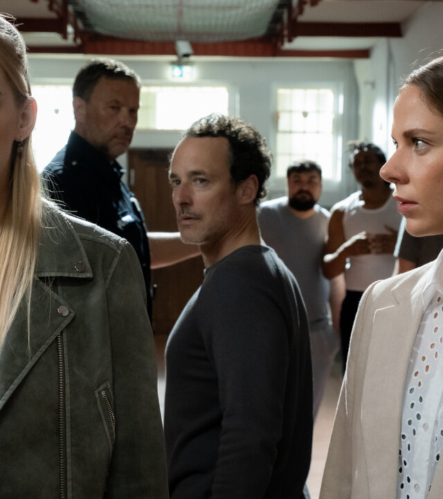 "Blutige Anfänger - Die Verwechslung": Auf dem Gang der JVA: Anastasia (Luise Emilie Tschersich) und Charlotte (Cheyenne Pahde) stehen im Vordergrund nebeneinander. Anastasia starrt geradeaus. Charlotte hat sich zu ihr gewandt und sieht sie von der Seite an. Hinter ihnen steht Juri (Sascha Tschorn). Er hat sich in Richtung Anastasia gedreht und sieht sie an. Neben ihm steht ein Justizvollzugsbeamter (Komparse). Im Hintergrund stehen zwei weitere Insassen (Komparsen).