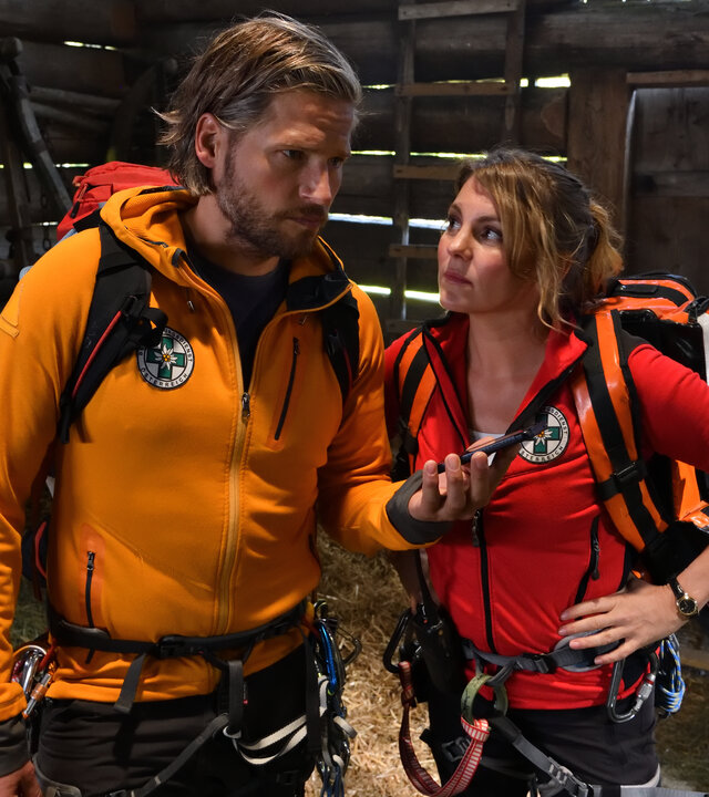 "Die Bergretter - Mutterglück (2)": In einer Scheune stehen die Bergretter Markus (Sebastian Ströbel) und Katharina (Luise Bähr) nebeneinander. Während Markus sein Handy in der Hand vor sie hält, schaut sie ihn fragend an.