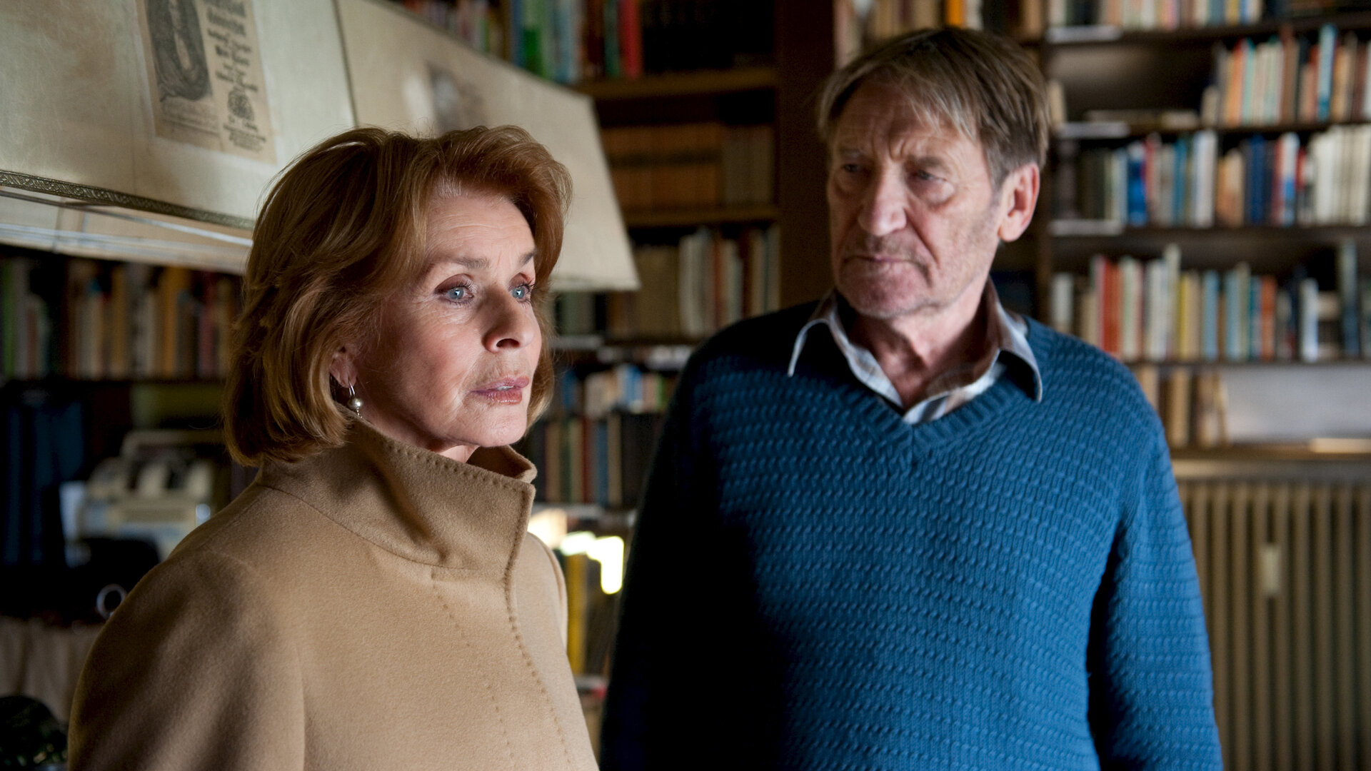 "Und alle haben geschwiegen": Paul (Matthias Habich) und Luisa (Senta Berger) stehen in einem Innenraum vor einem Bücherregal. Paul schaut zu Senta, die nachdenklich in die Ferne blickt.