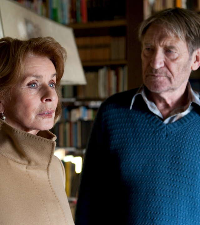 "Und alle haben geschwiegen": Paul (Matthias Habich) und Luisa (Senta Berger) stehen in einem Innenraum vor einem Bücherregal. Paul schaut zu Senta, die nachdenklich in die Ferne blickt.