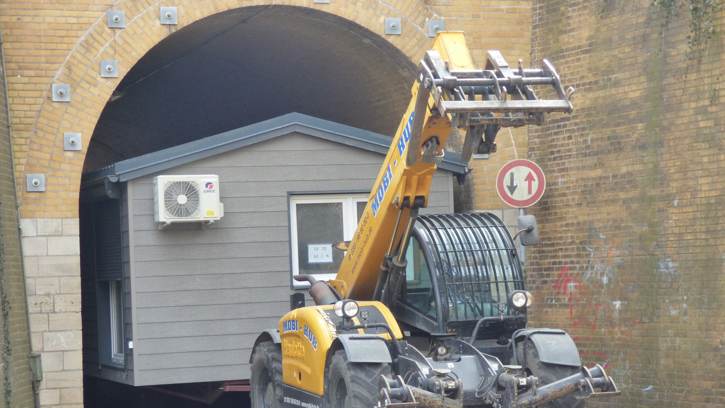 "Terra Xpress - Zu wenig, zu teuer - wird Wohnen zum Luxus?": Ein Bagger zieht ein Containerhaus durch einen Tunnel. Der Bagger ist bereits durch, das Haus noch nicht.