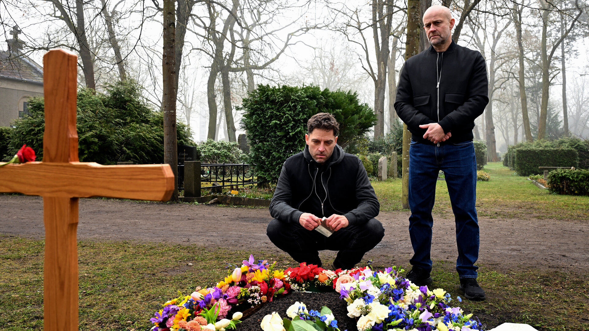 "Jenseits der Spree - Der Fall Andermatt": Levin Andermatt (Timur Isik) und Robert Heffler (JÃ¼rgen Vogel) befinden sich auf einem Friedhof an einem frisch geschmÃ¼ckten Grab. Levin Andermatt hockt vor dem Grab und blickt auf die Blumenarrangements, wÃ¤hrend Robert etwas dahinter steht und ebenfalls auf die GrabstÃ¤tte schaut. Am Grab steht ein hÃ¶lzernes Grabkreuz, hinter ihnen verlÃ¤uft ein Friedhofsweg.