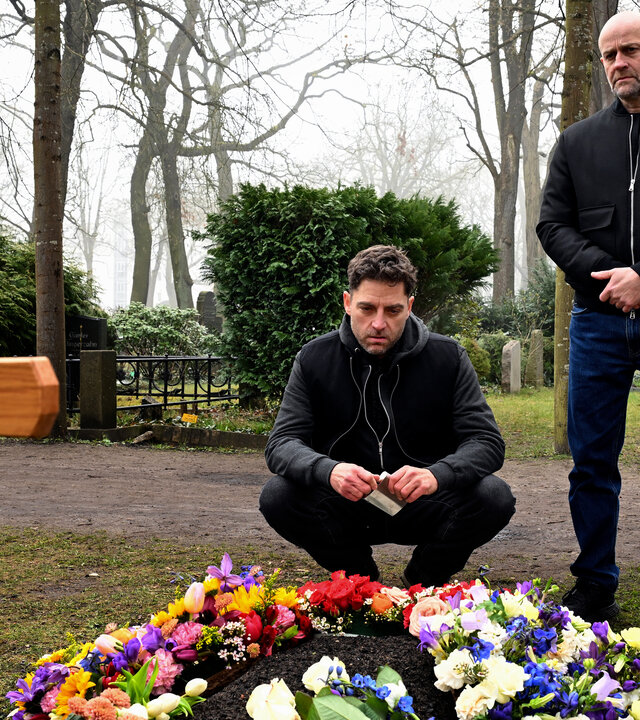 "Jenseits der Spree - Der Fall Andermatt": Levin Andermatt (Timur Isik) und Robert Heffler (JÃ¼rgen Vogel) befinden sich auf einem Friedhof an einem frisch geschmÃ¼ckten Grab. Levin Andermatt hockt vor dem Grab und blickt auf die Blumenarrangements, wÃ¤hrend Robert etwas dahinter steht und ebenfalls auf die GrabstÃ¤tte schaut. Am Grab steht ein hÃ¶lzernes Grabkreuz, hinter ihnen verlÃ¤uft ein Friedhofsweg.