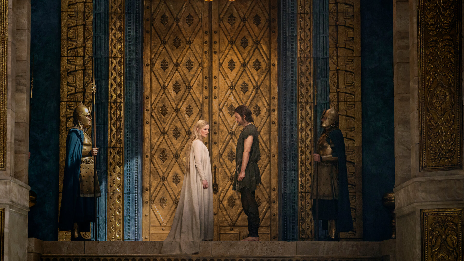 "Der Herr der Ringe: Die Ringe der Macht - Adar": Galadriel (Morfydd Clark) und Halbrand (Charlie Vickers) stehen sich in einem prunkvoll ausgestatteten Raum gegenüber. Über ihnen hängt ein Kronleuchter. Sie werden von zwei Wachen (Komparsen) beobachtet.
