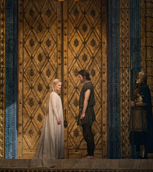 "Der Herr der Ringe: Die Ringe der Macht - Adar": Galadriel (Morfydd Clark) und Halbrand (Charlie Vickers) stehen sich in einem prunkvoll ausgestatteten Raum gegenüber. Über ihnen hängt ein Kronleuchter. Sie werden von zwei Wachen (Komparsen) beobachtet.