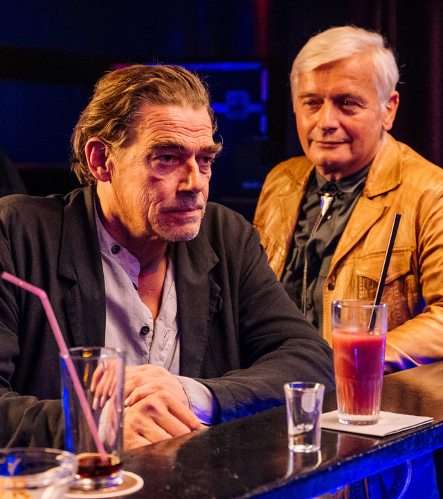 "SOKO Köln - Bukowski auf der Flucht": Viktor Kampe (Horst Kotterba) steht neben Fritz Bukowski (Martin Feifel) in einer Bar. Er hat einen Arm auf den Tresen gelegt und sich Bukowski lächelnd zugewandt. Bukowski hat sich mit beiden Armen auf dem Tresen abgestützt und blickt angespannt nach vorne. Vor ihm steht ein leeres Schnapsglas und ein roter Longdrink. Die Bar ist spärlich beleuchtet, die Theke wird durch blaues Licht angestrahlt. Im Hintergrund stehen zwei Security-Männer (Komparsen).