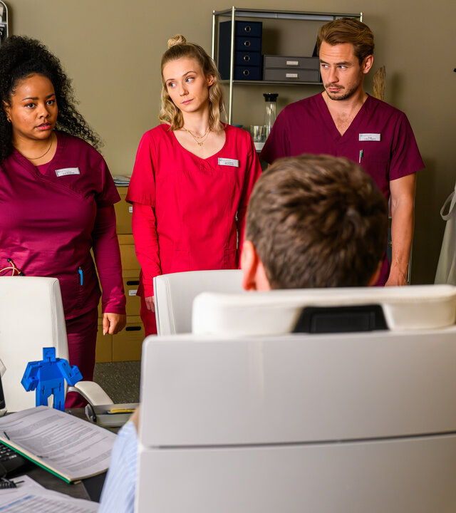 "Bettys Diagnose - Alle für eine": Betty (Henrike Hahn), Zoe (Catherine Chikosi), Mia (Antonia Döring), Lukas (Niklas Löffler) und Mechthild Puhl (Sybille J. Schedwill) stehen vor dem Schreibtisch von Dr. Walden (Constantin Lücke), der mit dem Rücken zur Kamera auf einem Stuhl sitzt. Auf dem Schreibtisch sind ein Monitor, ein Telefon und Papiere zu sehen. Im Hintergrund hängen zwei Arztkittel an einem Garderobenständer.